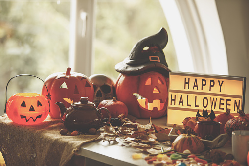 Front view of Happy Halloween elements placed on the table with decorative colorful pumpkins, dried leaves and tea pot against window in daylight.